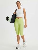 Women's lime green 14" high-waisted crossover capris with pockets, white tank top, and sneakers.