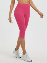 Woman wearing rose red high-waisted 17" capri leggings with two side pockets and white athletic shoes. #color_Rose Red