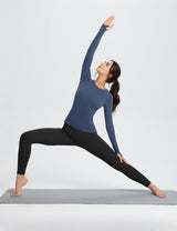 Woman in blue long-sleeve top and black leggings performing warrior yoga pose on gray mat.