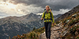 Realistic blonde woman wearing waterproof hiking pants walking on a rugged mountain trail.