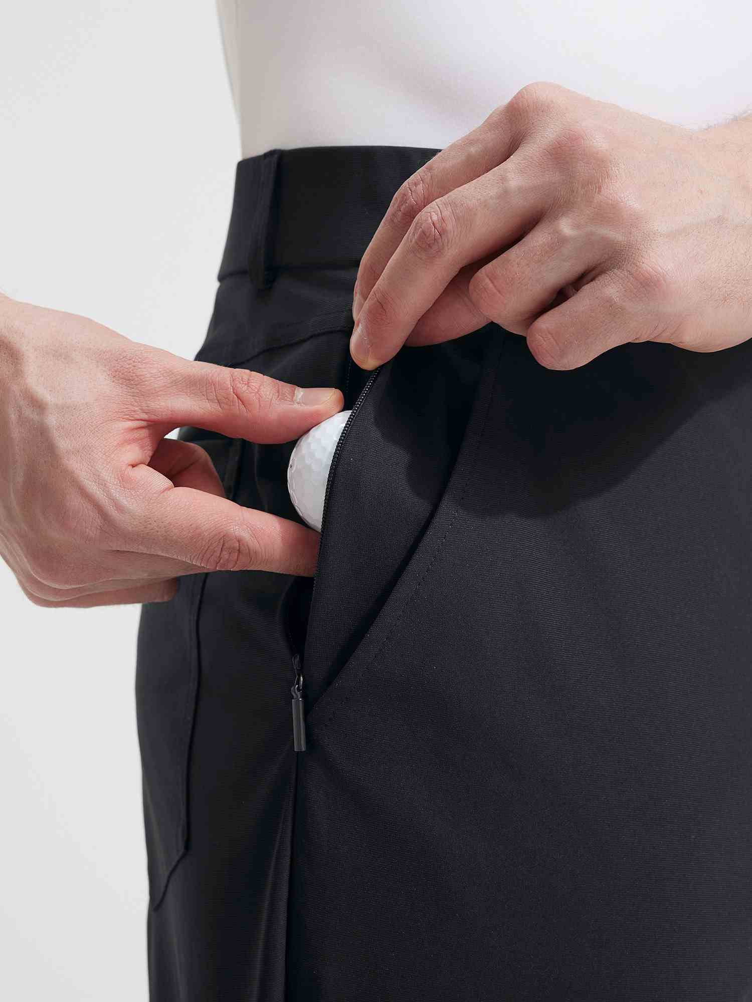 Close-up of black Baleaf golf pants, hand placing golf ball into zippered side pocket. #color_Black