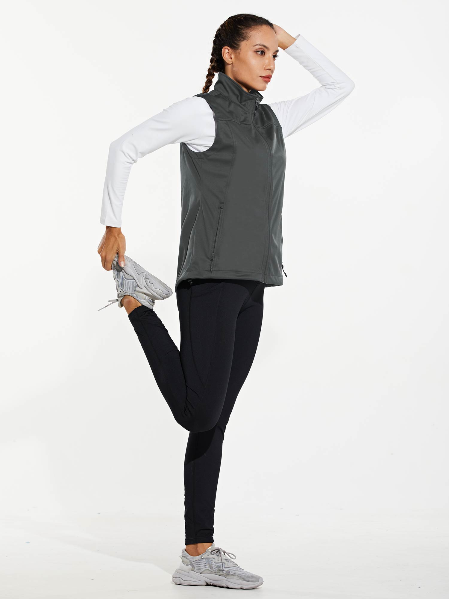 Gray windproof softshell vest on woman with white shirt, black leggings, & sneakers, performing a stretch. #activewear #vest