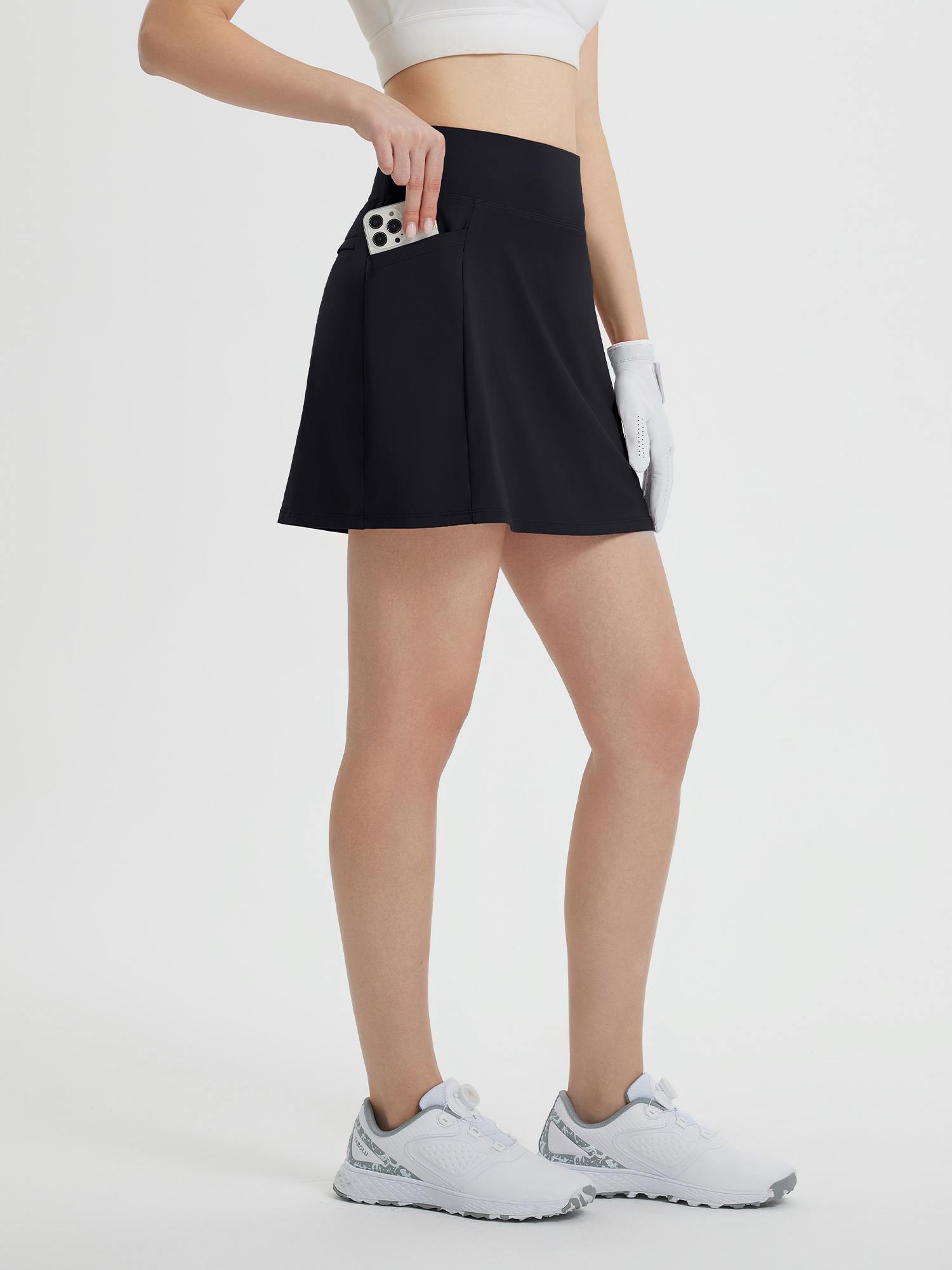 Black high-waisted golf skort with pockets holding phone, golf glove, white shoes. #color_Black