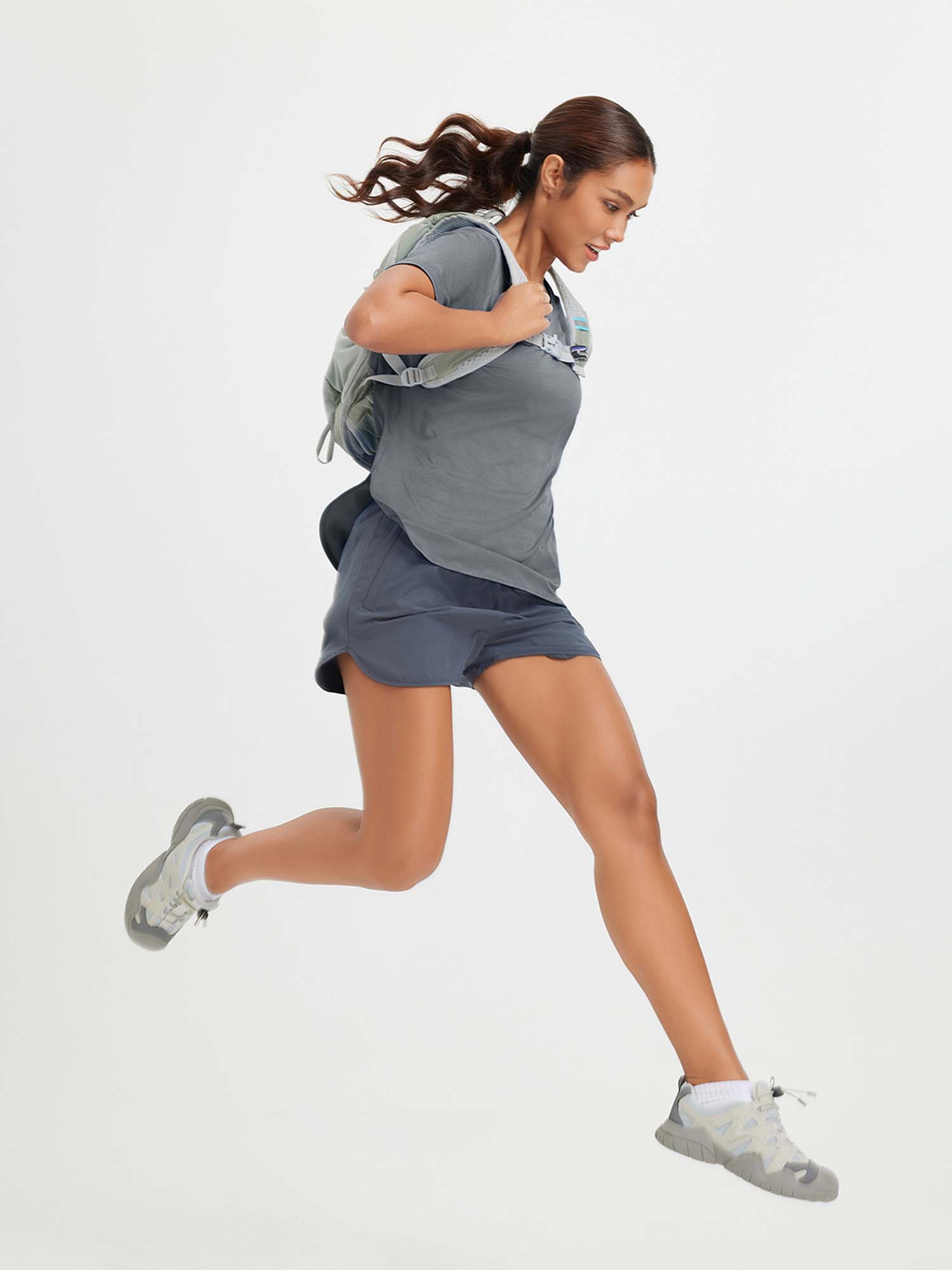Woman in grey t-shirt and blue shorts running, wearing a backpack and athletic shoes. #color_Heather Light Gray