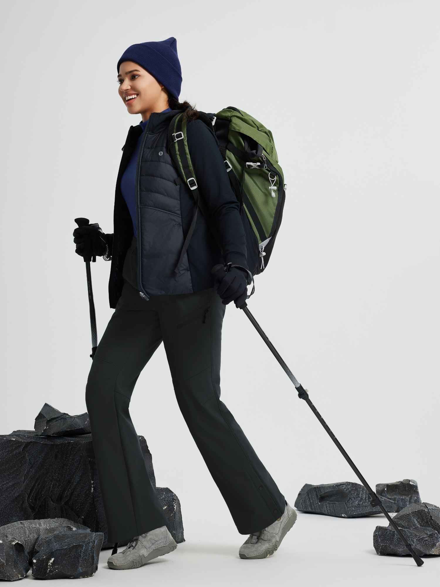 Woman hiking in dark softshell straight-leg pants, green backpack, trekking poles.
