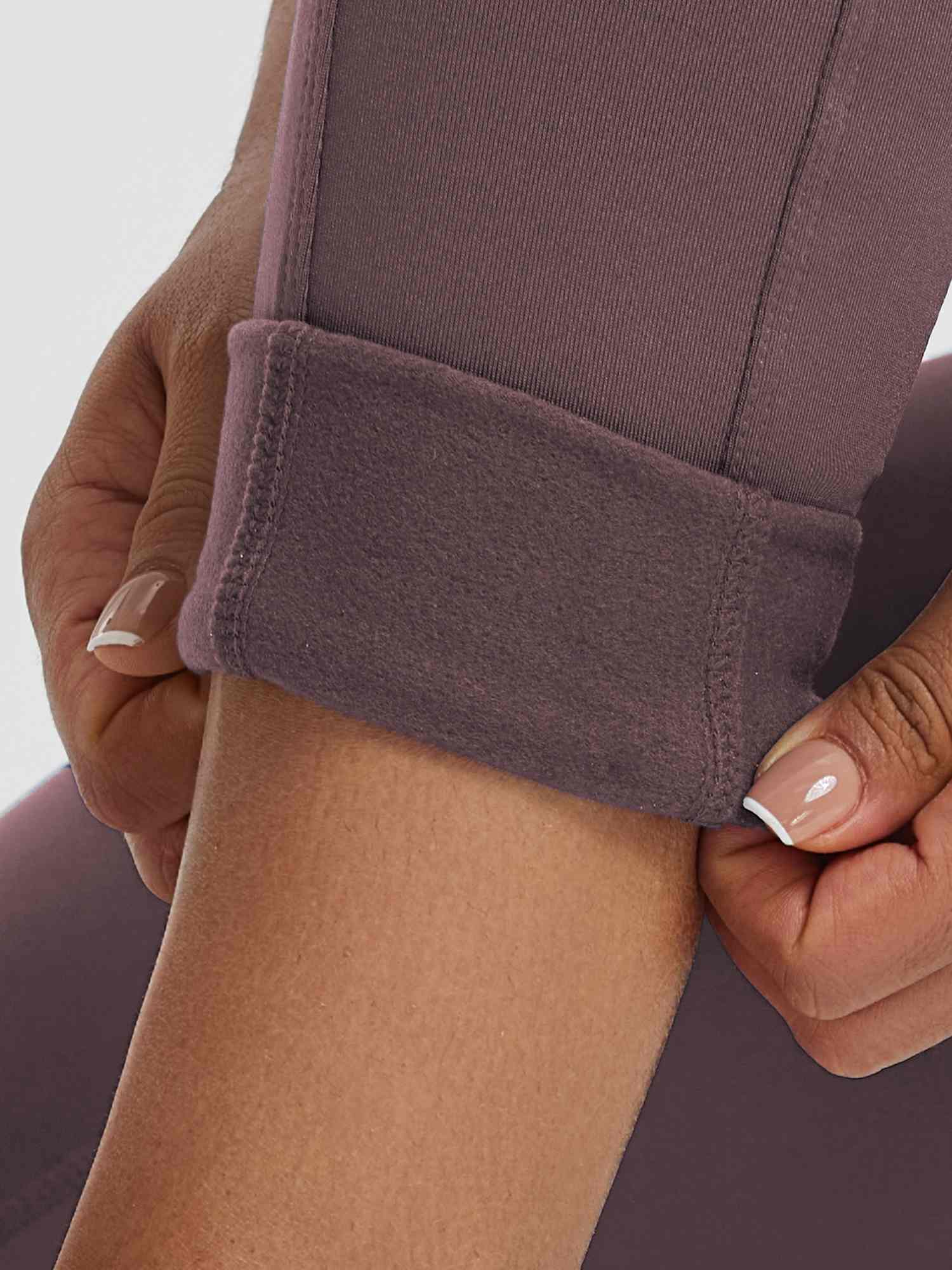 Close-up: Hands rolling mauve high-waisted fleece running leggings hem. #color_Dark Brown