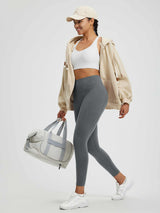Model in grey high-waisted fleece yoga leggings, beige hoodie, and cap, holding a duffel bag. #color_Heather Dark Gray