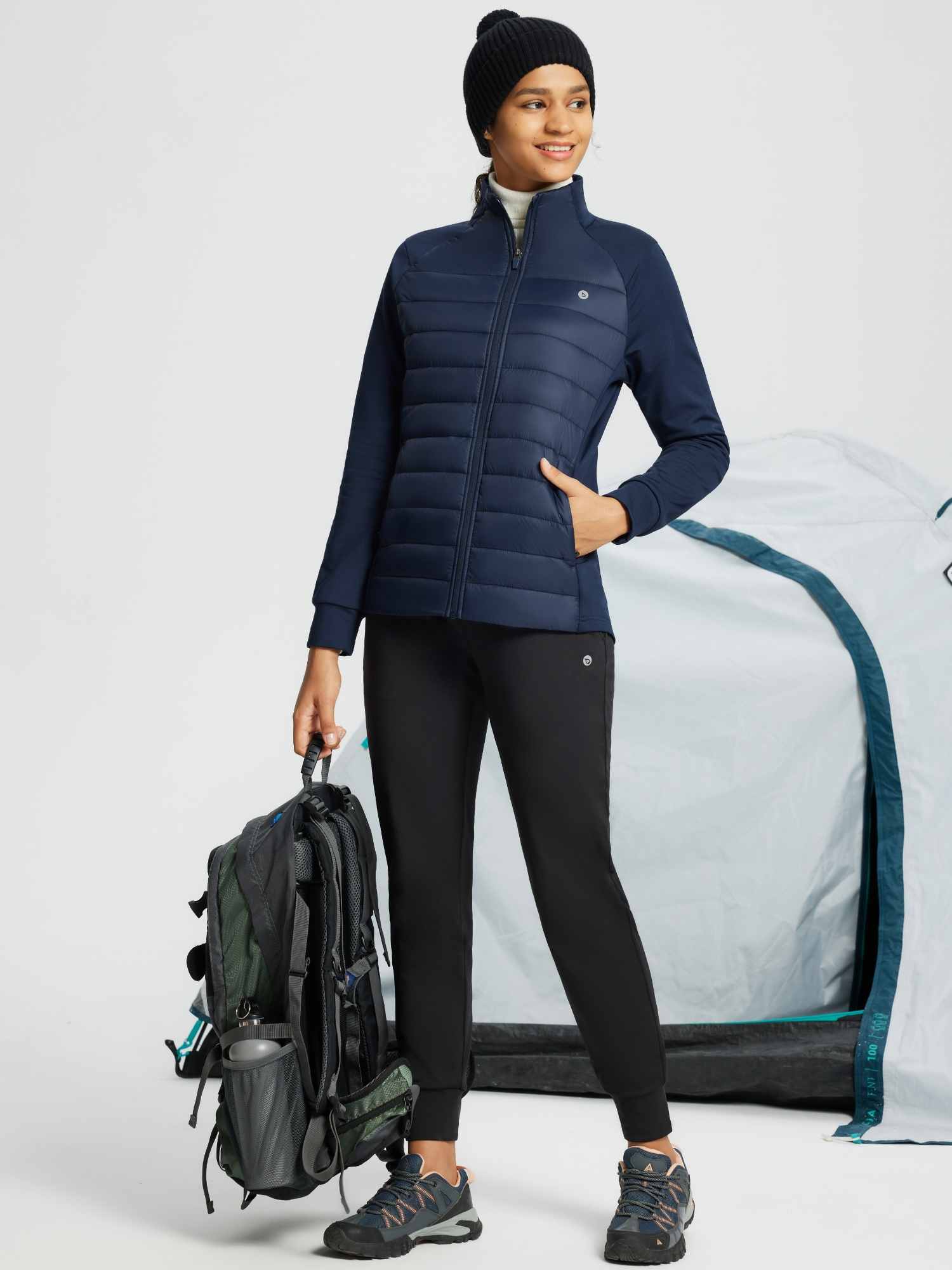 Women's navy quilted outdoor jacket with stretch sleeves, holding backpack near tent. #color_Navy Blue