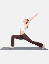 Woman in white sports bra and brown bootcut workleisure pants performing a warrior yoga pose on a grey mat.