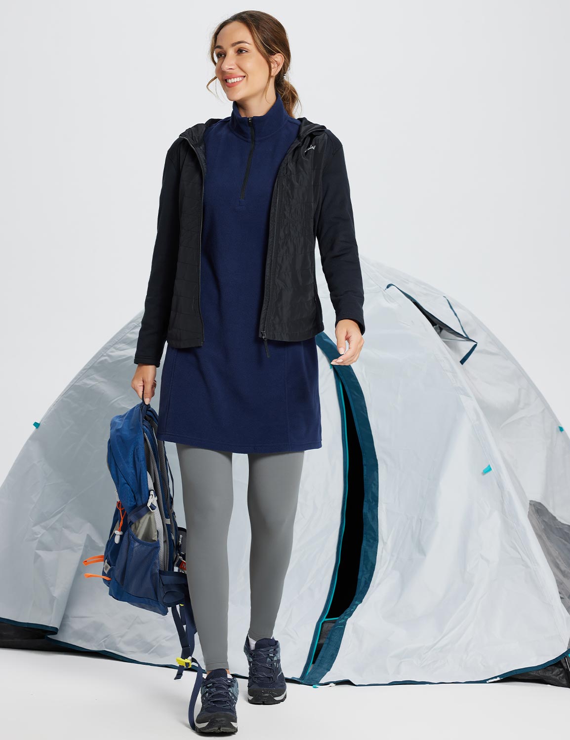 Woman in navy thermal tunic dress, black jacket, grey leggings, and hiking boots holding a backpack. #color_Navy Blue